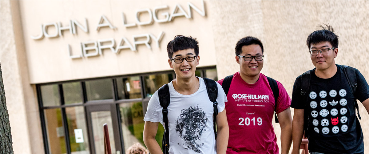Three male international students walking and smiling outside Logan Library on a sunny day.